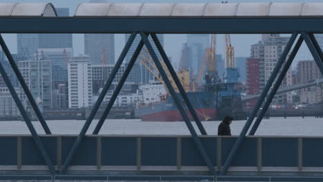 man walking over bridge with river thames industry, cargo ship and financial district in background