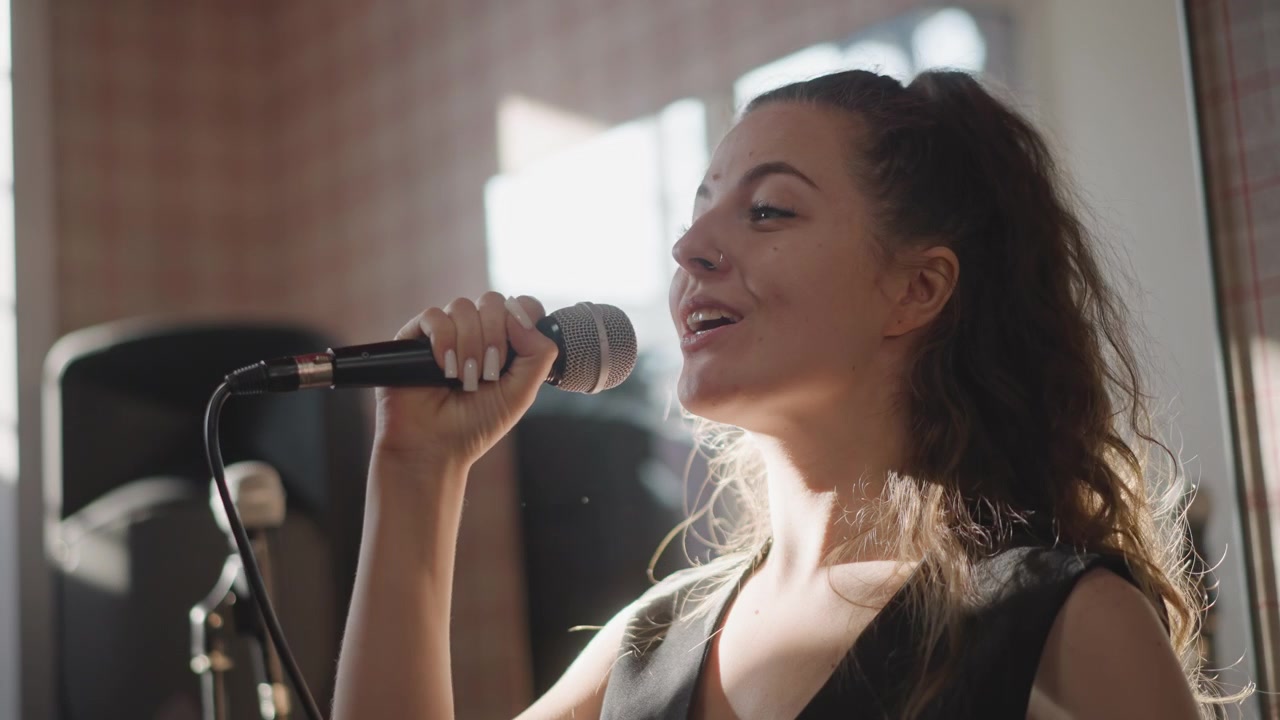 Singer smiling during vocal practice session, expressive phrasing and bright natural light, confident mic technique, upbeat mood with band equipment in background, musician delivering warm performance