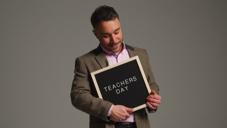 studio portrait of mature male teacher standing against grey background holding notice board reading teachers day