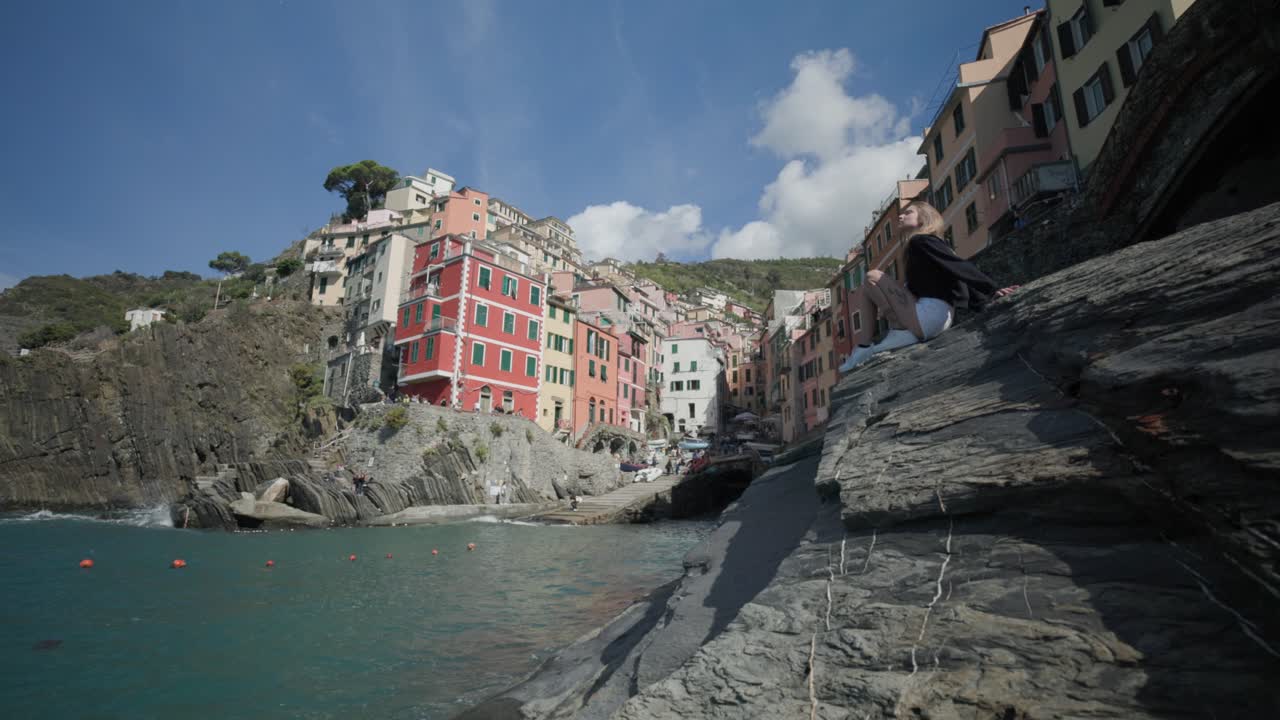 Touriste assise sur un rocher au bord de la mer, avec Riomaggiore en arrière-plan.