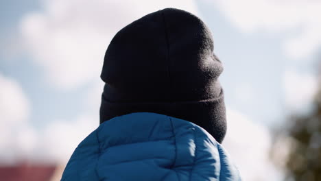 rear head view of individual wearing black beanie and blue puffer jacket, looking outdoors, the background shows a soft blue sky and blurred autumn foliage