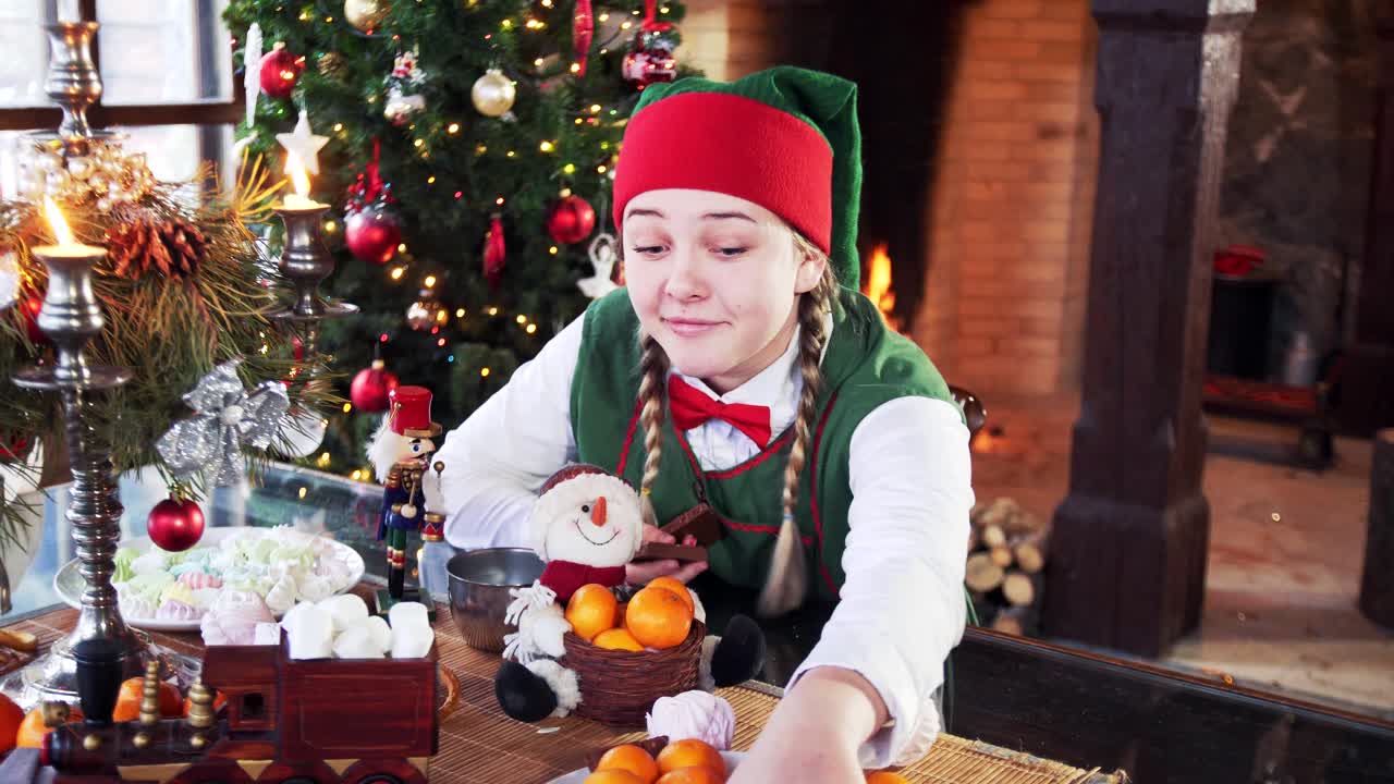 Girl in elf costume sitting at festive table. Funny elf takes many chocolate pieces from plate in Christmas decorated room