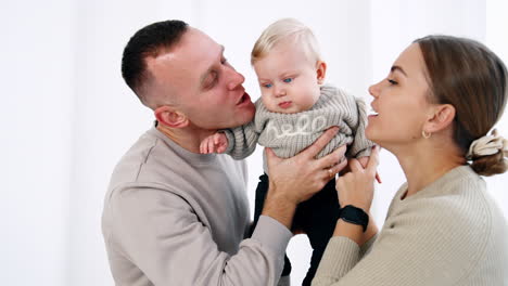 young family of three in grey sweaters. caucasian parents hold and kiss their infant son on the cheeks. white backdrop.
