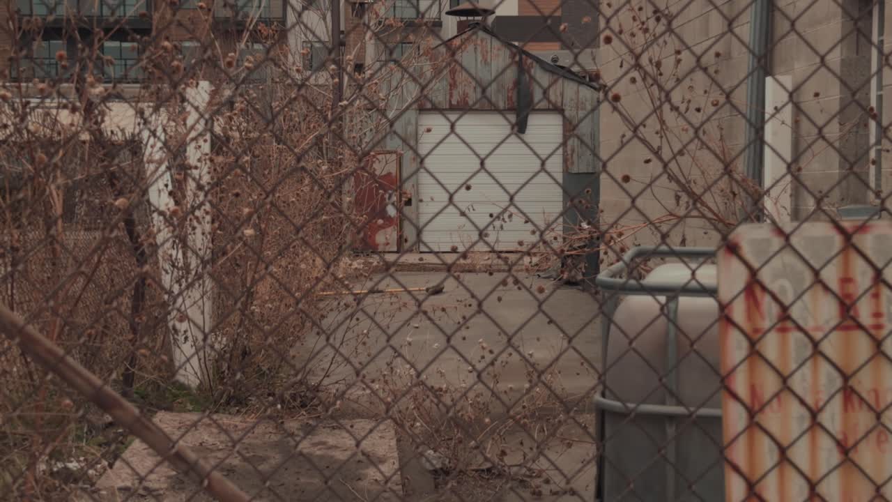 Slow steady zoom through chain link fence toward an abandoned industrial alley and loading area