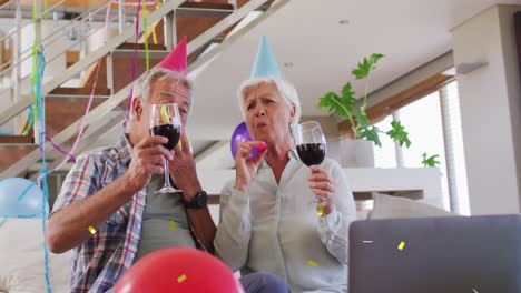 happy caucasian senior couple in party hats making birthday laptop video call drinking wine