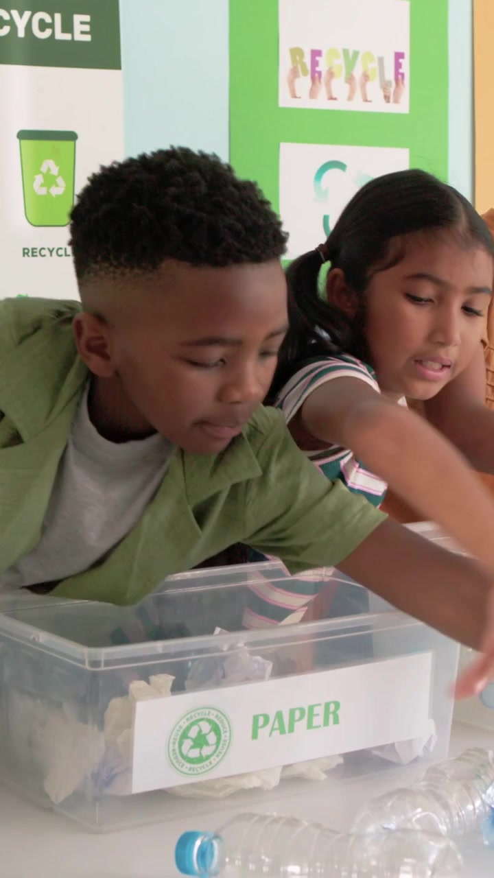 Vertical video: Adult offering recyclables, girl in green tee sorting paper into PAPER bin in class