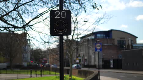 Radar-speed-sign-in-the-UK,-twenty-miles-per-hour,-happy-smiley-face-as-road-users-respect-speed-limit,-cars-and-scooters-driving-in-slow-motion-on-a-bright-sunny-day