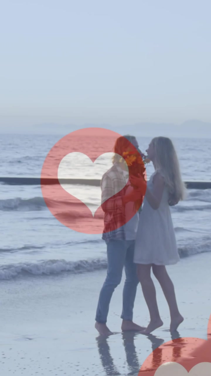 Plajda öpüşen beyaz tenli bir çiftin üzerinde süzülen birden fazla kırmızı kalp simgesi