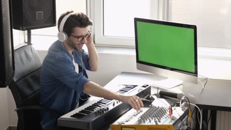 young man in sound recording studio. cheerful positive soundman using mixing console and listening music through headphones. green digital screen.