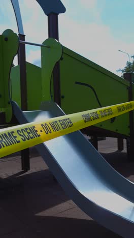 yellow and vibrant colored crime scene do not enter tape ribbon pulled in front of a green childrens playground that has a silde during a sunny day indicating a crime has happened, investigation