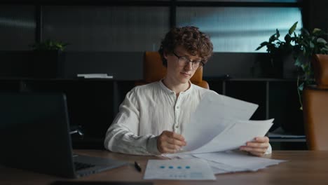 A-confident-young-guy-with-curly-hair-in-glasses-and-a-white-shirt-goes-through-documents-and-examines-papers-while-sitting-at-a-table-in-the-office