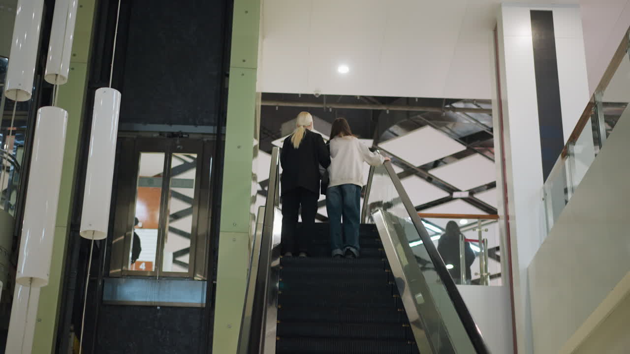 Back view of two ladies riding escalator up in modern shopping mall, casual clothing, relaxed moment highlighting friendship and lifestyle atmosphere inside contemporary retail environment