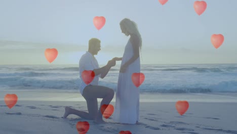 multiple heart balloons floating against man proposing a woman with a ring on beach