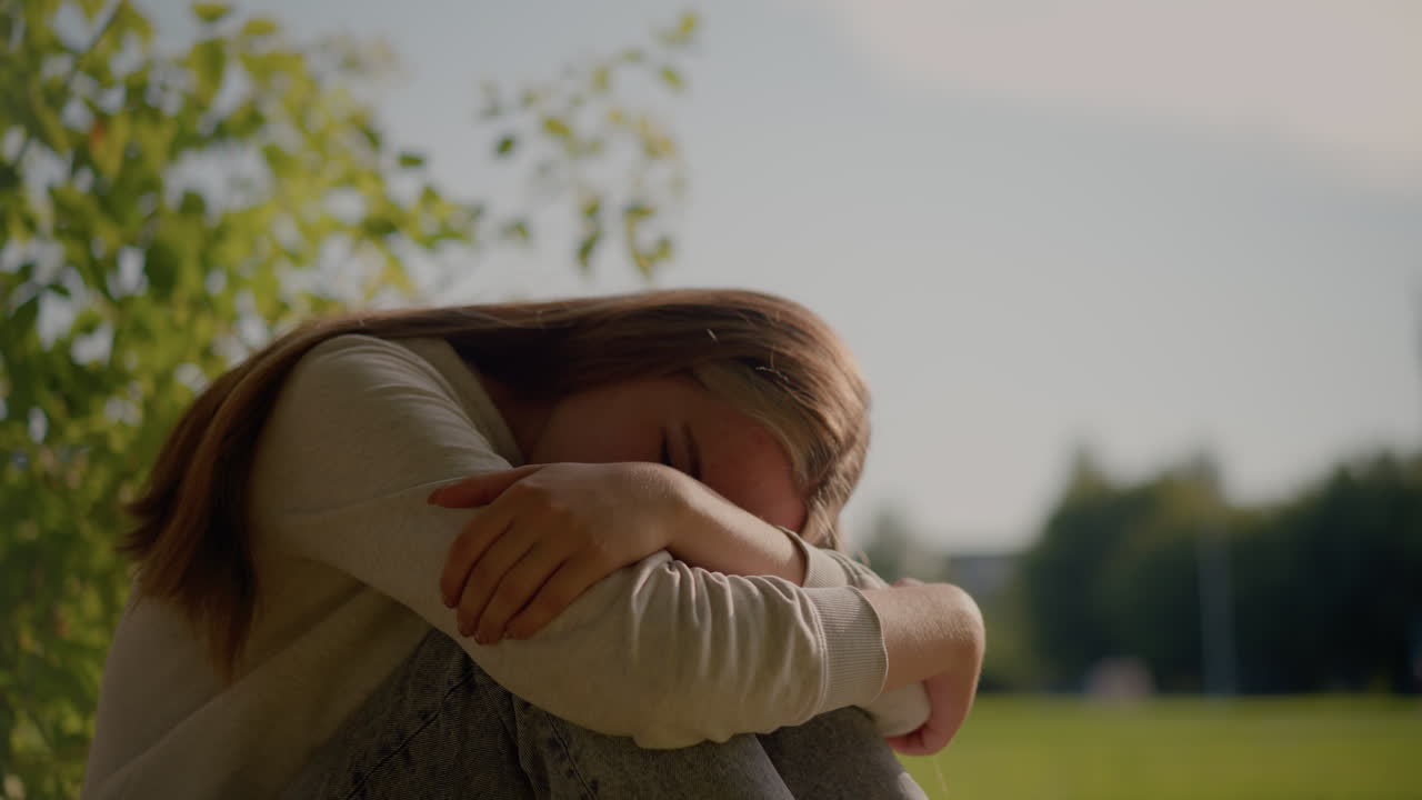 una joven pensativa se sienta en el suelo cubierto de hierba con los brazos cruzados, apoyando la cabeza en su mano mientras la luz del sol ilumina suavemente su rostro, rodeada de vegetación y fondo borroso
