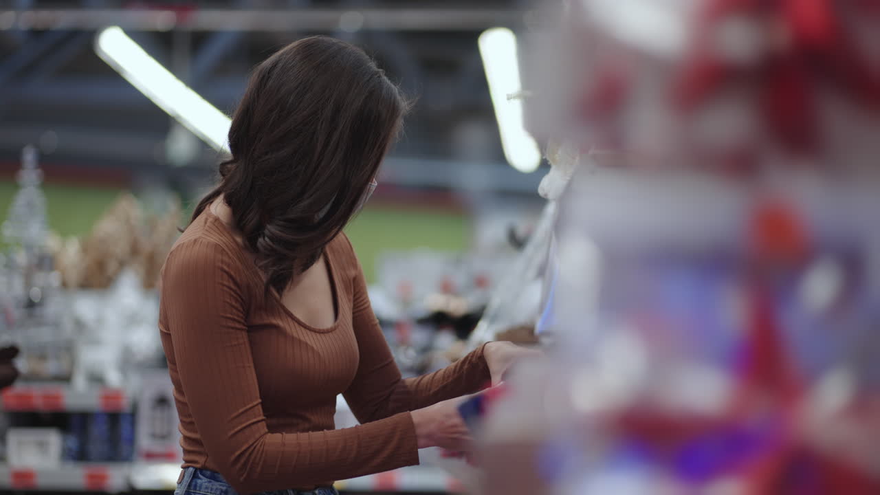 una mujer joven con una máscara recoge una pelota de navidad roja antes de navidad. elige decoraciones navideñas. preparándose para la navidad en la pandemia de coronavirus. guirnaldas
