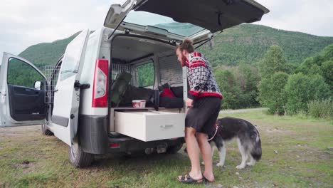 Joven-Organizando-Su-Ropa-En-El-Cajón-De-La-Furgoneta-Con-Malamute-De-Alaska