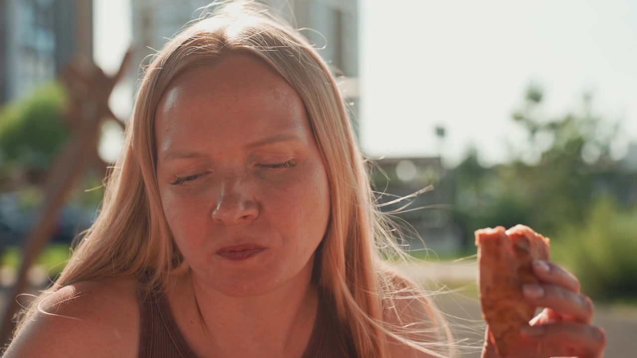Una donna con lunghi capelli biondi tiene in mano e mangia un pezzo di cibo all'aperto alla luce del giorno. Lo sfondo mostra edifici con vegetazione sfocata e una luce solare intensa che crea ombre morbide sulla sua pelle.