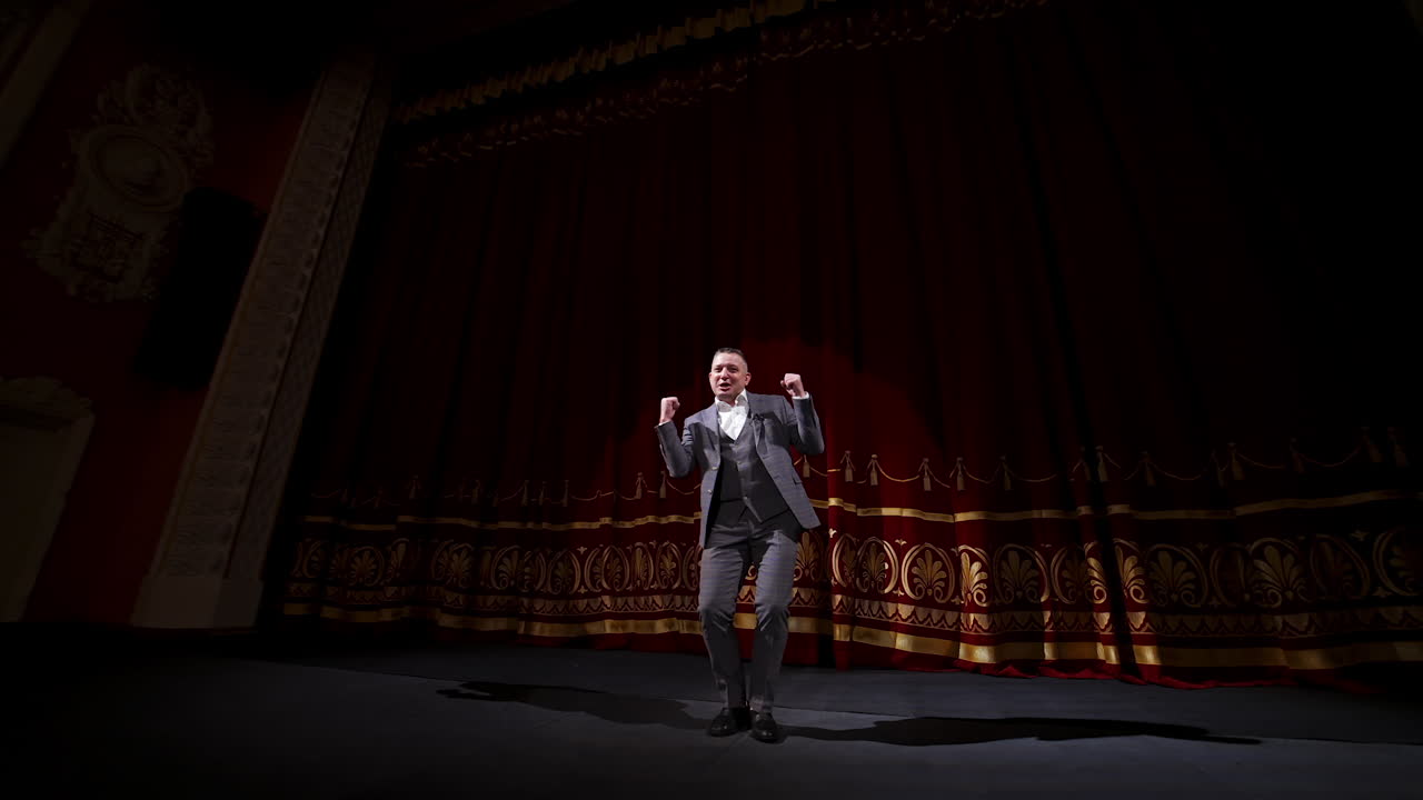 Actor in suit on large theater stage. Male comedian telling jokes and dancing gestures on the scene. Joyful performer on stage.