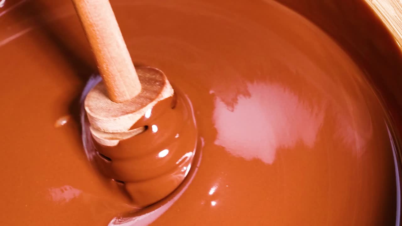 Un bastoncino di legno si immerge in cioccolato liscio e lucido, creando increspature e riflessi nel ricco liquido