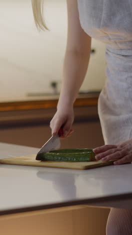 young wife cuts cucumber on wooden cutting board cooking vegetarian salad for dinner on table with bowl in modern kitchen close view slow motion