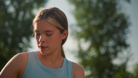 young lady in light blue top adjusts posture while focusing downward with hair tied back, with background features blurred view of green leaves