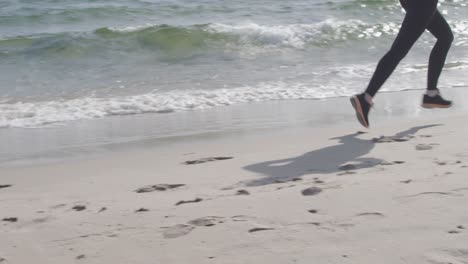 Kamerafahrt-Mit-Den-Beinen-Einer-Sportlerin-In-Sportbekleidung,-Die-In-Zeitlupe-Entlang-Der-Wasserlinie-Am-Strand-Joggt