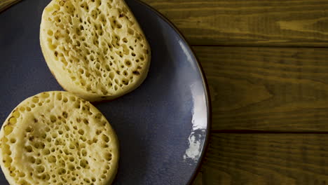 top down shot of a male placing some tasty and delicious crumpets on a blue stone plate on a wooden background, buttering them then taking a bite and placing back down