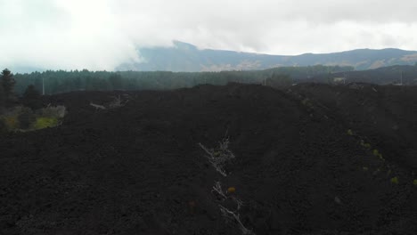 AERIAL-DOLLY-IN:-Drone-flying-over-a-valley-of-lava-rocks,-in-Mount-Etna,-Sicily,-on-a-foggy-day,-revealing-a-desolate-and-martian-look-panorama-with-the-top-of-the-crater-in-the-distance