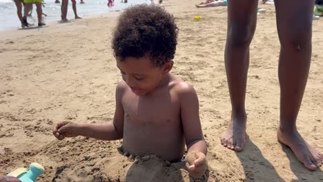 4-year-old black kid buried waist down playing with the sand on the beach next to his sister. he is wearing black curly hair. handheld camera. 4k