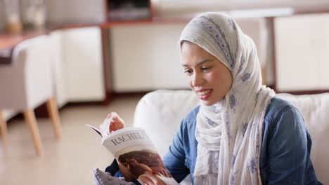 Happy-biracial-woman-in-hijab-reading-book-on-sofa-at-home-with-copy-space,-slow-motion