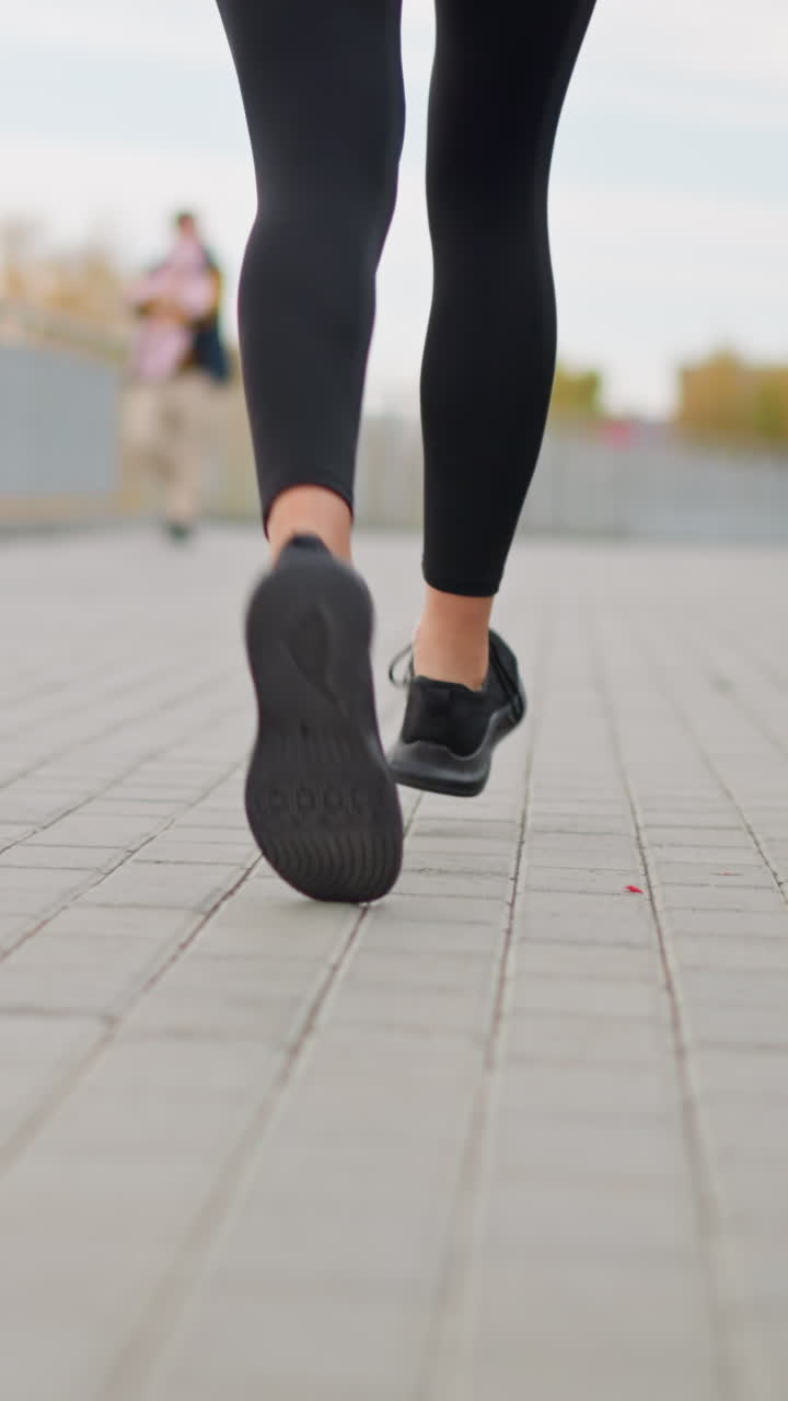 Close-up view of athlete woman running on pavement in black leggings and sneakers, with two women passing in blurred background, promoting active lifestyle and fitness