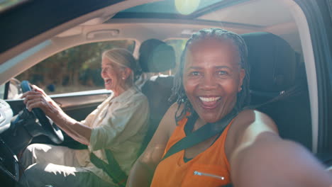 passenger taking selfie on mobile phone as two senior female friends enjoy day trip out in car