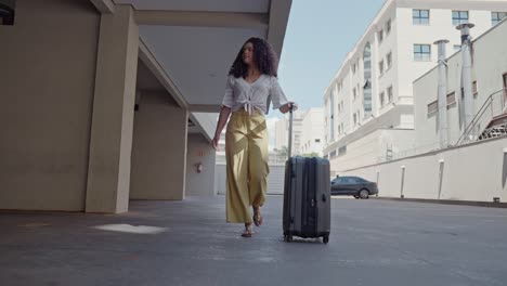 young latin girl tourist walks with a suitcase on wheels around city on hotel garage background a modern building. brazilian female traveling woman student walking the hotel.