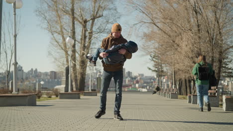 a front view of a father carrying his child while walking down a paved street, with a woman walking in the opposite direction, with a blur view of city behind him