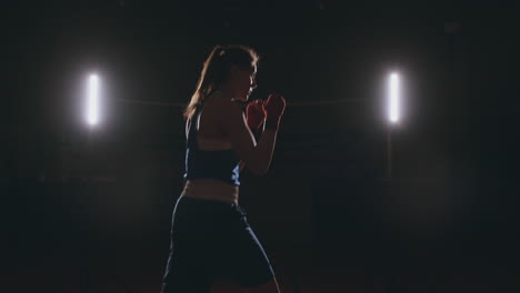 a beautiful sports boxer woman in red bandages on her hands and a blue t-shirt is fighting with a shadow practicing the speed and technique of punches. camera movement side view. steadicam shot. preparing for self-defense