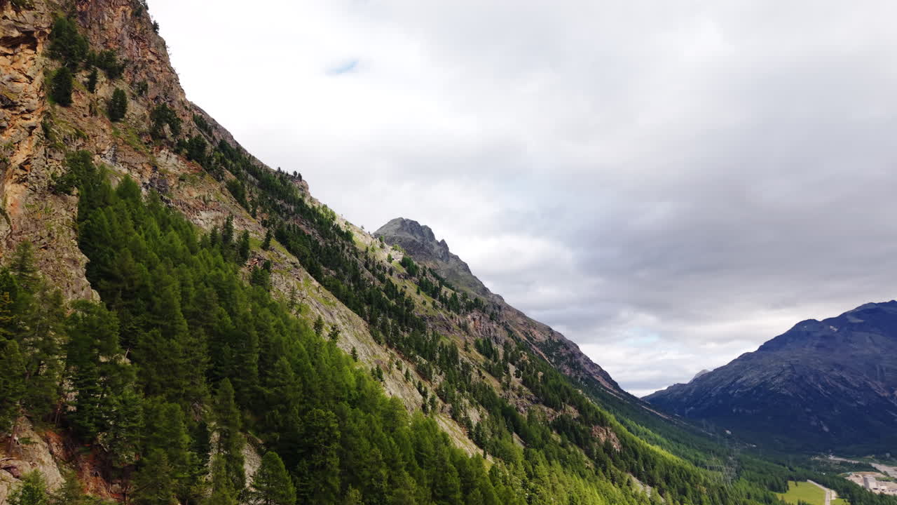 Forward drone flight along forested mountain slope toward distant Swiss alpine peaks