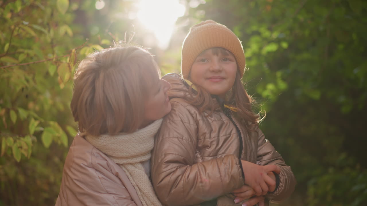 mother holding smiling child close in sunlit autumn woods, gently touching child head as warm backlight halo glows around, both wrapped in knit scarf and jackets, expressions tender and joyful