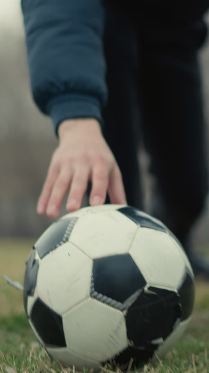 Blurred view of a coach dropping a soccer ball on a grassy field in a stadium as two people begin to play, set against a background view of buildings and trees
