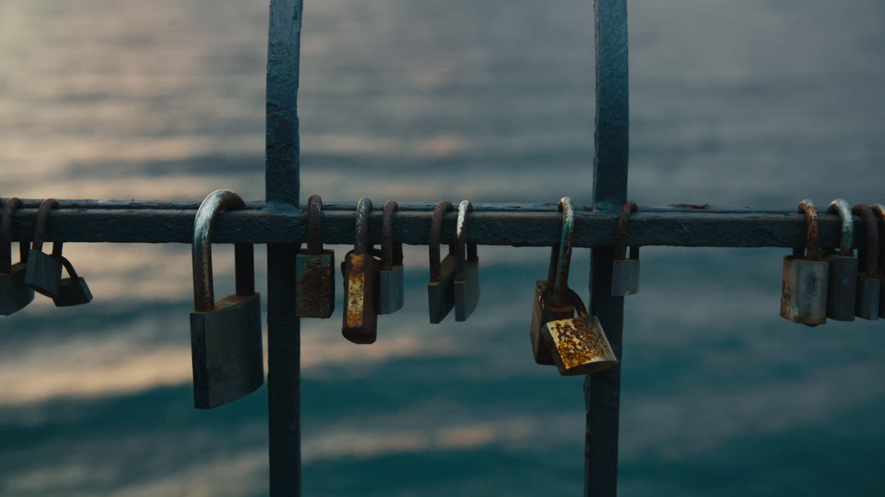 Close-up of rusted and worn love padlocks hanging from a seaside iron fence, calm ocean blurred in the background under soft evening light