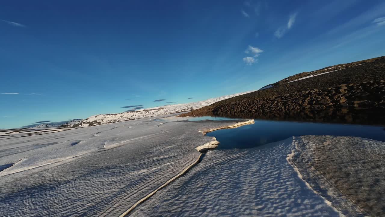 Low FPV drone flight along the icy edge of Heiðarvatn dam in eastern Iceland, following open water beside frozen lake surfaces under clear skies near Seydisfjordur