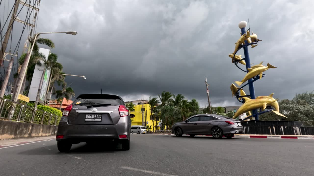 Car approaches large yellow sculpture on city street, dramatic rain clouds overhead, daytime, steady camera