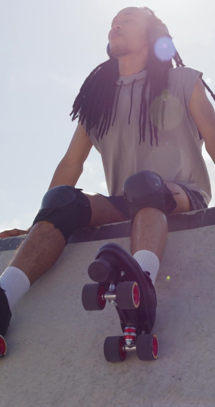 Young Man With Dreadlocks Sitting on Skate Park Ramp Wearing Roller Skates