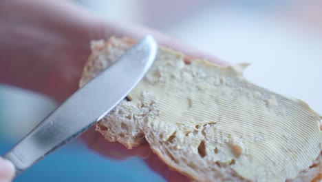 Primer-Plano-De-Una-Mano-Esparciendo-Mantequilla-Sobre-Una-Rebanada-De-Pan,-Enfoque-Suave,-En-Cámara-Lenta