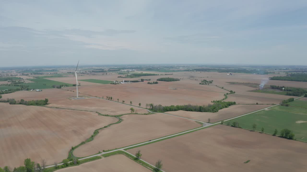 Wide aerial view of flat terrain, farm fields and large wind power turbine towering over the landscape