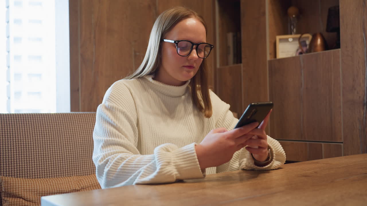 Side view of student wearing white sweater and glasses sitting at wooden desk while chatting on smartphone in cozy interior with warm lighting and books in background