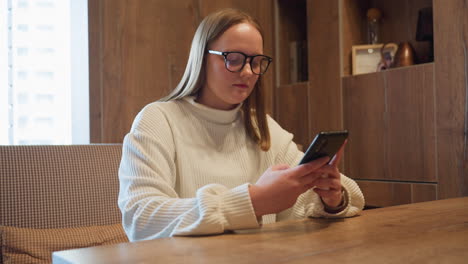 side view of student wearing white sweater and glasses sitting at wooden desk while chatting on smartphone in cozy interior with warm lighting and books in background