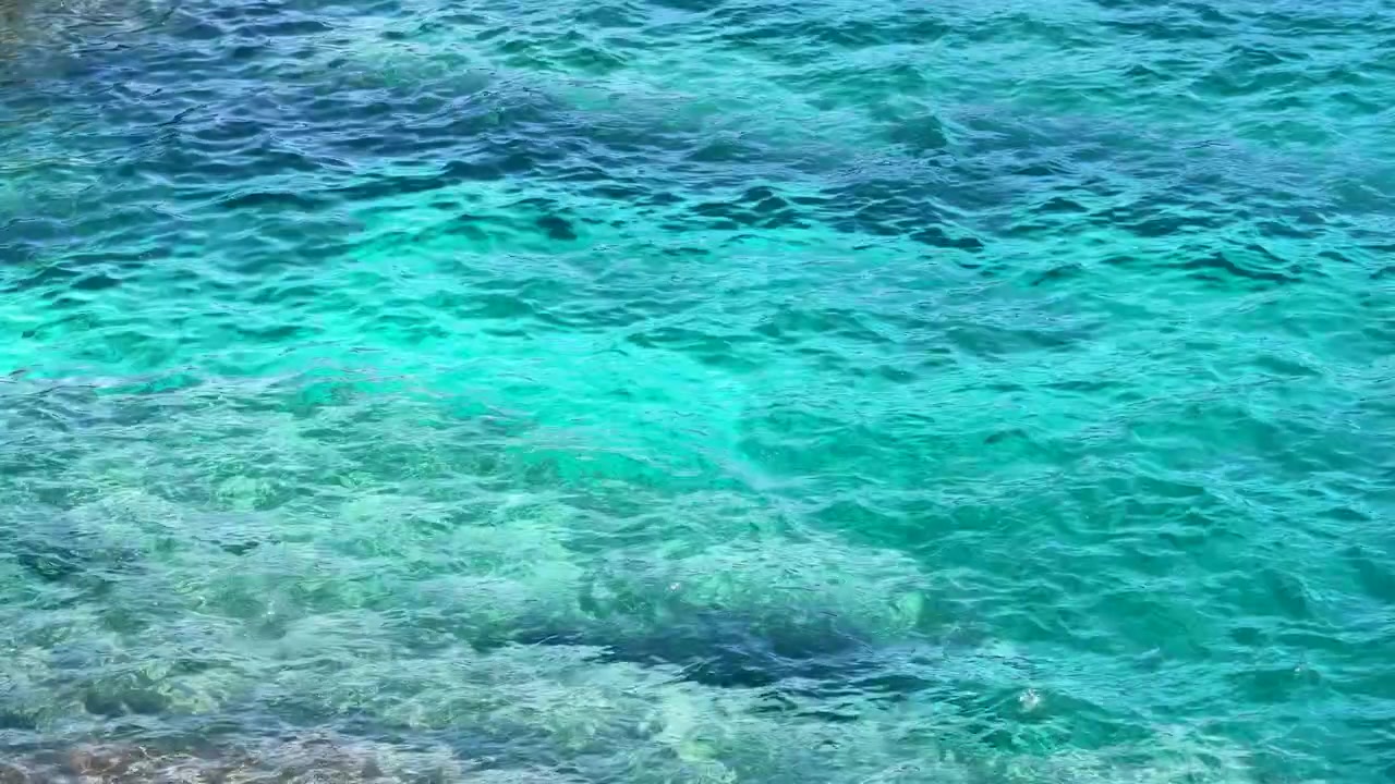 Imagem estática em ângulo alto de águas mediterrâneas turquesa cintilantes com leito marinho rochoso visível abaixo.