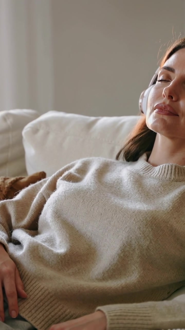 Vertikal video: Lyttende kvinde i beige sweater læner sig tilbage på sofaen, med hovedtelefoner på, blødt lys beroliger