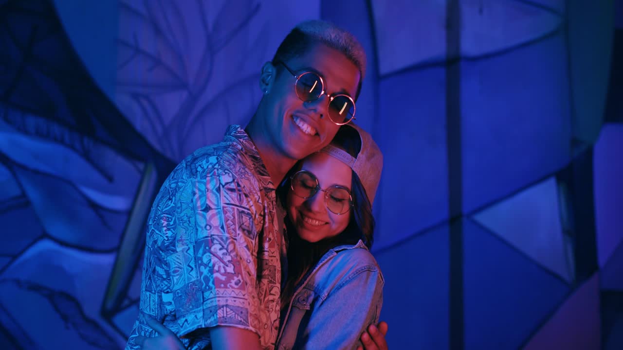 elegante pareja hipster multiétnica enamorada abrazándose y mirando la cámara al aire libre por la noche