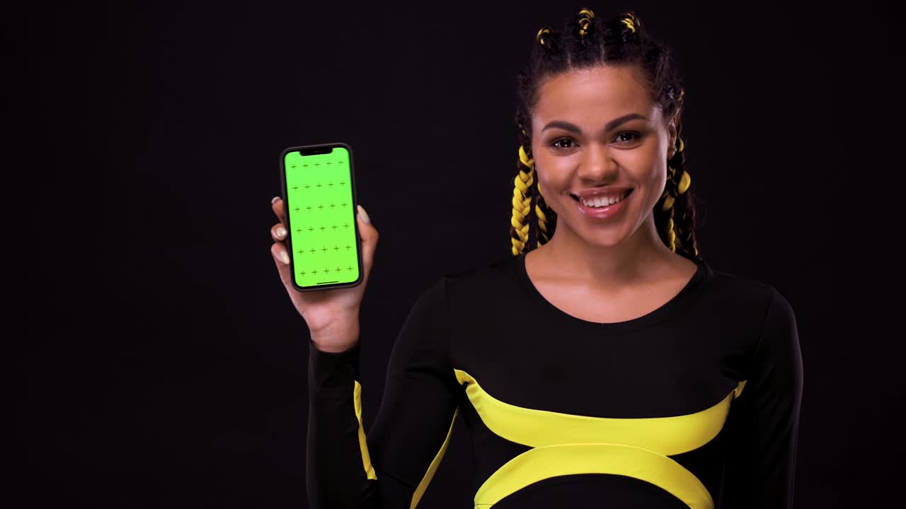 retrato de una chica afroamericana sonriente apuntando con el dedo a la pantalla del teléfono móvil con una llave cromática. mujer aislada sobre un fondo negro